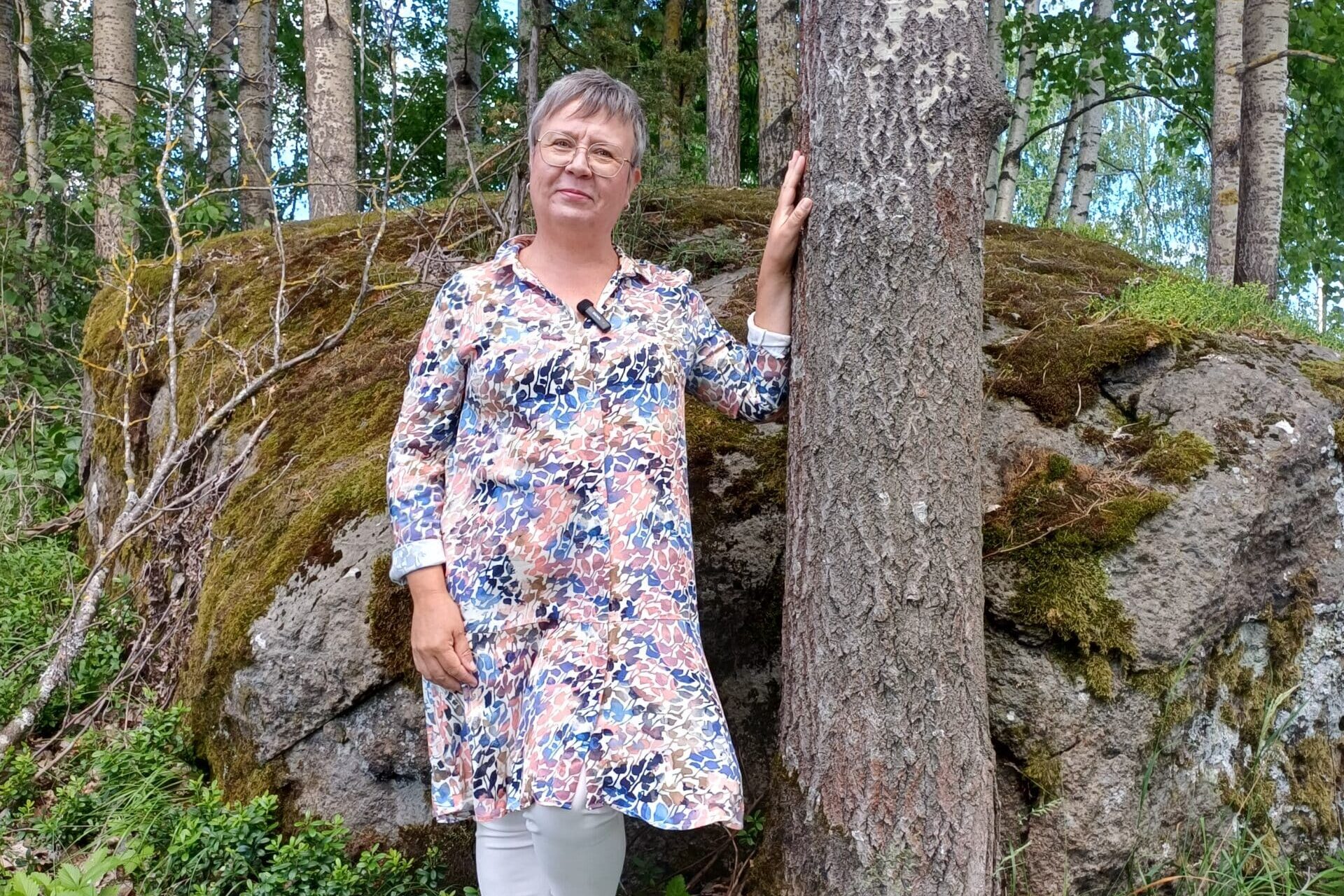 Kokemustoimija Marjo seisoo luonnossa ja nojailee puuhun.