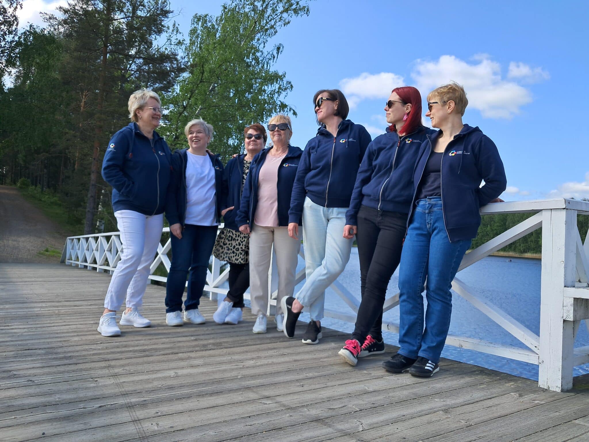 Seitsemän naista seisoo sillalla, Valkoisen sillankaiteen takana sininen järvi.